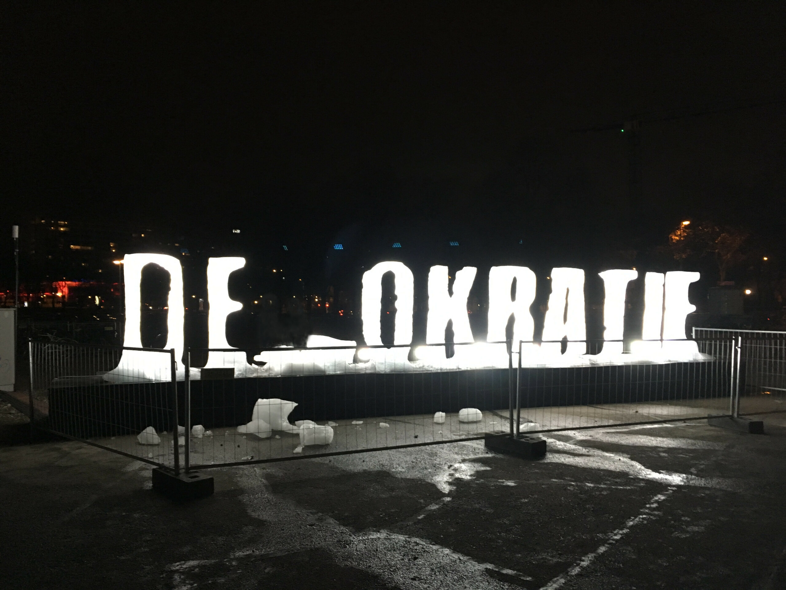 Foto des Schriftzugs Demokratie: Die Buchstaben aus Eis geformt, das M liebt zerbrochen daneben, so dass zu lesen ist: "DE OKRATIE"